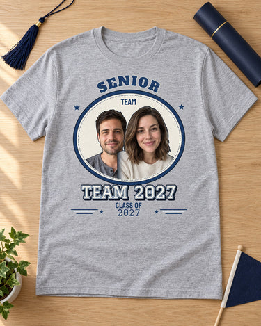 Sport grey TEAM 2027 senior photo shirt laid on an oak desk with a graduation tassel, diploma tube and navy pennant, showing two portraits.