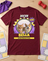 Maroon Bella pet superhero tee with a purple cape laid on pale wood beside a plain toy cape, rubber dog toy and comic pages with empty panels.