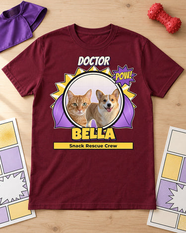 Maroon Bella pet superhero tee with a purple cape laid on pale wood beside a plain toy cape, rubber dog toy and comic pages with empty panels.