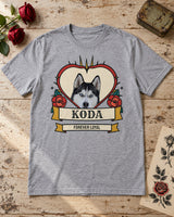 Sport grey Koda tattoo pet tee laid on pale wood beside red roses, a vintage tin and botanical tattoo flash, with a red heart and teal leaves.