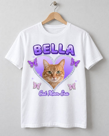White Y2K pet photo t-shirt on a wooden hanger with an orange tabby cat in a lilac rhinestone heart, glittery butterflies and the name Bella