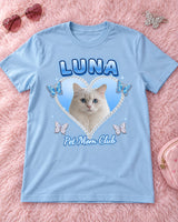 Light blue Y2K pet photo t-shirt laid flat on a fluffy pink rug with heart sunglasses, a hair clip and a makeup bag, a white cat in a baby blue heart with the name Luna