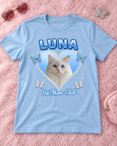 Light blue Y2K pet photo t-shirt laid flat on a fluffy pink rug with heart sunglasses, a hair clip and a makeup bag, a white cat in a baby blue heart with the name Luna